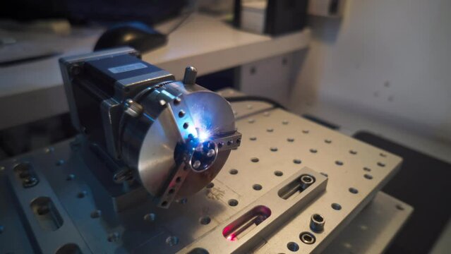 Desktop device for laser engraving rings rotating in process of work, camera passing by. Workshop space, computer mouse on background, metal work surface equipped with fasteners, laser beam movement.
