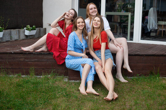 Group of young smiling gorgeous barefoot women sitting on wooden veranda near green grass, looking at camera, laughing.