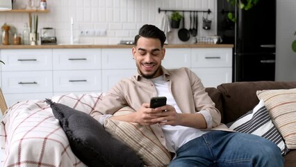Attractive indian or arabian young man, sit in a modern living room on comfortable sofa, uses his smartphone, texting message, browsing social media, looks away, smiles. The concept of using gadgets