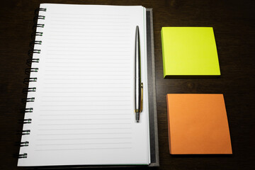 A Pen is on the notebook and beside the Sticky note on the wooden table.