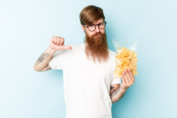 Young caucasian man holding a bag of chips isolated on blue background feels proud and self...