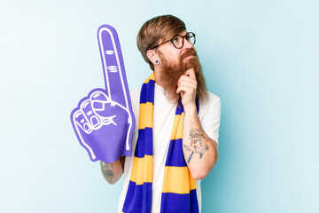 Young sport fan holding a foam hand isolated on blue background looking sideways with doubtful and skeptical expression.