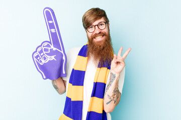 Young sport fan holding a foam hand isolated on blue background joyful and carefree showing a peace symbol with fingers.