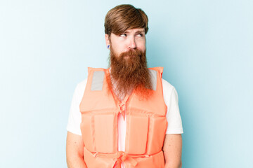 Young caucasian man wearing life jacket isolated on blue background confused, feels doubtful and...