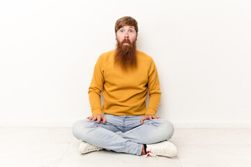 Young caucasian man sitting on the floor isolated on white background shrugs shoulders and open eyes confused.