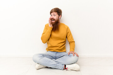 Young caucasian man sitting on the floor isolated on white background is saying a secret hot braking news and looking aside