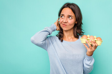 Young hispanic woman holding eggs isolated on blue background touching back of head, thinking and...