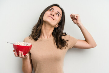 Young caucasian woman holding bowl of cereals isolated on white background raising fist after a...