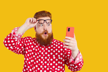 Mobile offer. Funny chubby bearded man with emotional shocked expression looks at screen of mobile phone, raising his glasses. Surprised fat man in red shirt with smart phone on yellow background. - obrazy, fototapety, plakaty
