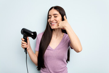 Young caucasian woman holding hairdryer isolated on blue background showing a mobile phone call gesture with fingers.