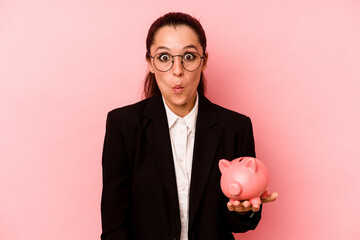 Young business caucasian woman holding piggybank isolated on pink background shrugs shoulders and...