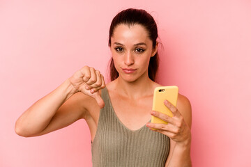 Young caucasian woman holding mobile phone isolated on pink background showing a dislike gesture, thumbs down. Disagreement concept.