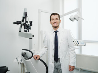 Naklejka na ściany i meble Portrait of happy male dentist wearing lab coat in white dental clinic