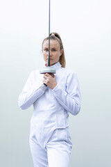 Caucasian woman in white fencing costume hold epee in front of face, wears jacket, pants. Sports,...
