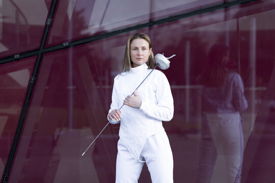 Portrait Of White Female Fencer Posing On Red Glass Background, Wears Professional Costume: White Jacket, Pants, Put Epee On Shoulder. Expensive Sport, Experienced Coach, Healthy Lifestyle.Horizontal.