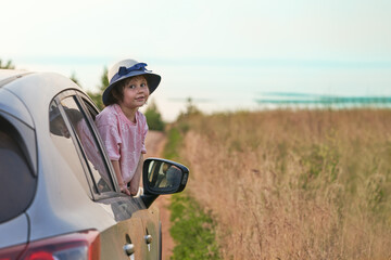 A happy little girl in a hat in the car window at the entrance to the seashore. Selective focus.