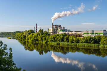 pipes of woodworking enterprise plant sawmill with beautiful reflection in blue water of river. Air...