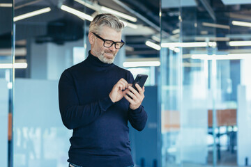 Senior gray-haired businessman in office using phone, happy investor reading message and smiling,...