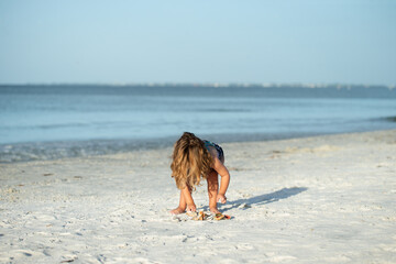 Little girl with long hair in her face bent over collecting shells in a pile on a sandy beach under...