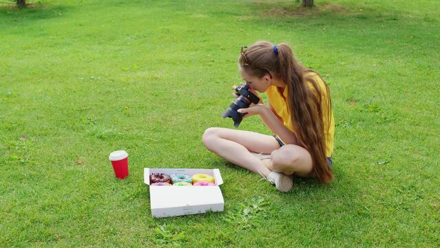 Female food photographer sits on grass, takes pictures of donuts inside paper packaging. Girl shooting doughnuts with professional camera in box on lawn for her blog about nutrition, diets, fast food