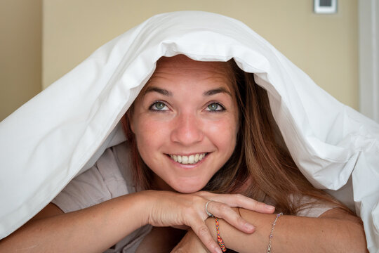 Mujer sonrriente bajo s&aacute;banas blancas pensando algo lindo luego de levantarse