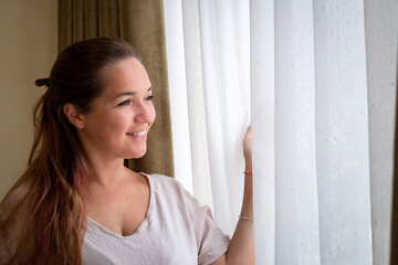 Mujer sonriente mirando por la ventana de su habitación perdida en sus pensamientos