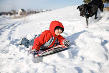 A boy sledding in the winter time with a black lab dog