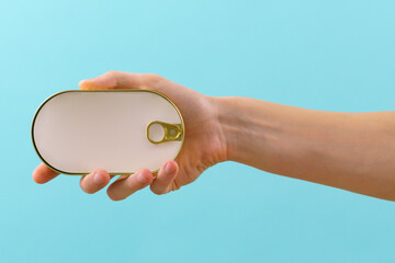 The man's hand holds a closed golden rounded empty fish tin can with an empty white paper mock-up on a blue background. Sardine. Container. Can. Fish. Package. Ring. Nutrition. Closed. Canning. Close
