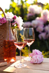 Bottle and glasses of rose wine near beautiful peonies on wooden table in garden