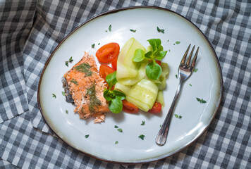 Light fish salad with baked salmon flesh, slices of cherry tomatoes, and cucumber slices, sprinkled with fresh herbs and garnished with corns salad leaves, top view, handmade plate