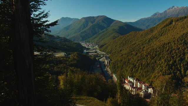 Breathtaking view of rosa khutor village among the majestic caucasus mountain range. clear sky with no clouds and sun rays falling down the forested mountains painted in autumn colours.