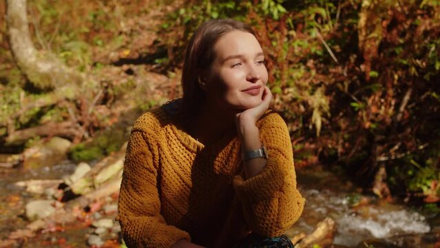 Portrait of pretty girl enjoing the beauty of autumn forest with flowing brook against the background. young woman is relaxing and smiling among the scenic fall foliage and plants.