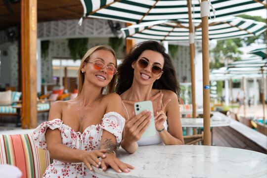 Two young beautiful smiling hipster female in trendy summer sundress.Taking photo selfie on mobile phone at outdoor cafe. Positive models having fun
