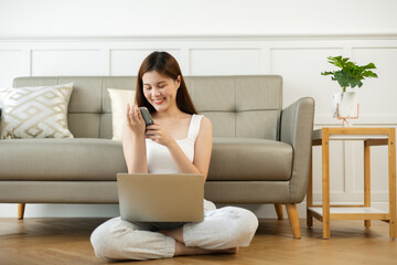 Freelance asian woman in glasses with mobile phone typing at laptop and working from home office. Happy girl sitting on couch in living room.