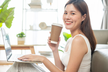 Freelance asian woman in glasses with mobile phone typing at laptop and working from home office. Happy girl sitting on couch in living room.