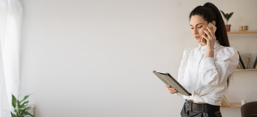 Elegant asian businesswoman using mobile phone after working on digital tablet, standing in office interior, woman manager talking to client using smartphone
