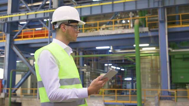 Engineer working in industrial factory.
The engineer in a white helmet and safety glasses is working inside the factory with his tablet in hand.
