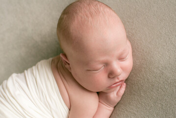 A cute baby sleeps wrapped in an airy milk-colored blanket on a beige plaid on a bed at home.