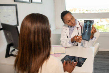 Confident doctor showing x-ray picture of lungs and explaining diagnosis to patient. Young African American pulmonologist consulting woman in clinic. Medical consultation, pulmonology concept - obrazy, fototapety, plakaty