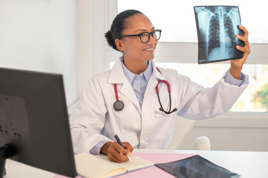 Portrait of happy female doctor looking at x-ray image and making notes in notebook. Young African American pulmonologist working at table in her office. Pulmonology concept
