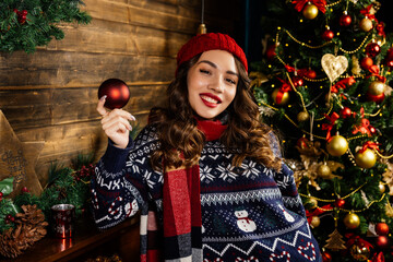 A beautiful brunette stands by the fireplace against the backdrop of a Christmas tree and Christmas decorations.