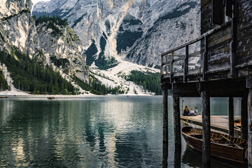 Young people sitting on boardwalk and enjoy amazing view of Braies Lake with the hut and boats in...