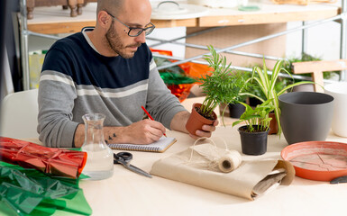 seated man writing plant data which he is holding with his hand. person surrounded with gardening products for the home and floral decoration
