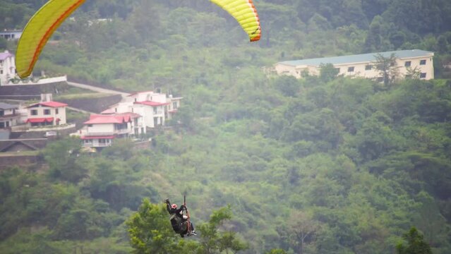 zoomed shot showing para gliding people with a bright orange yellow glider landing in front of buildings in the middle of hills mountains near nanital bir billing showing a popular adventure sport for