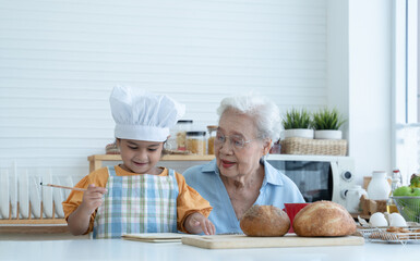 Asian family grandmother and grandchild have fun cooking at home kitchen together, little cute granddaughter with chef hat and apron is writing down recipe and how to make bread or cookie from Grandma