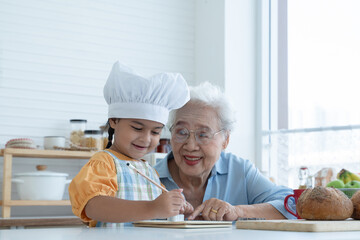 Asian family grandmother and grandchild have fun cooking at home kitchen together, little cute granddaughter with chef hat and apron is writing down recipe and how to make bread or cookie from Grandma - obrazy, fototapety, plakaty