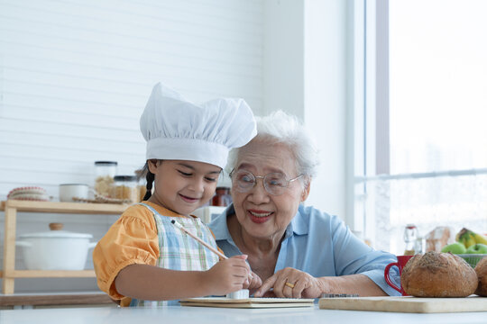 Asian Family Grandmother And Grandchild Have Fun Cooking At Home Kitchen Together, Little Cute Granddaughter With Chef Hat And Apron Is Writing Down Recipe And How To Make Bread Or Cookie From Grandma