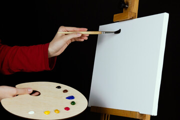 Female hand in red sweater with artist brush and paint palette near white empty canvas on wooden professional easel. Suitable as a mockup for your pictures and inscriptions.