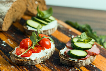 Three kinds of sandwiches with cottage cheese and vegetables: cucumbers, tomatoes, basil and arugula on dark bread dusted with flour on a wooden kitchen dividing board. For websites, menus, banners