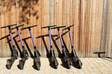 Row of electric scooters parked along wooden wall. Self-service street transport rental service.